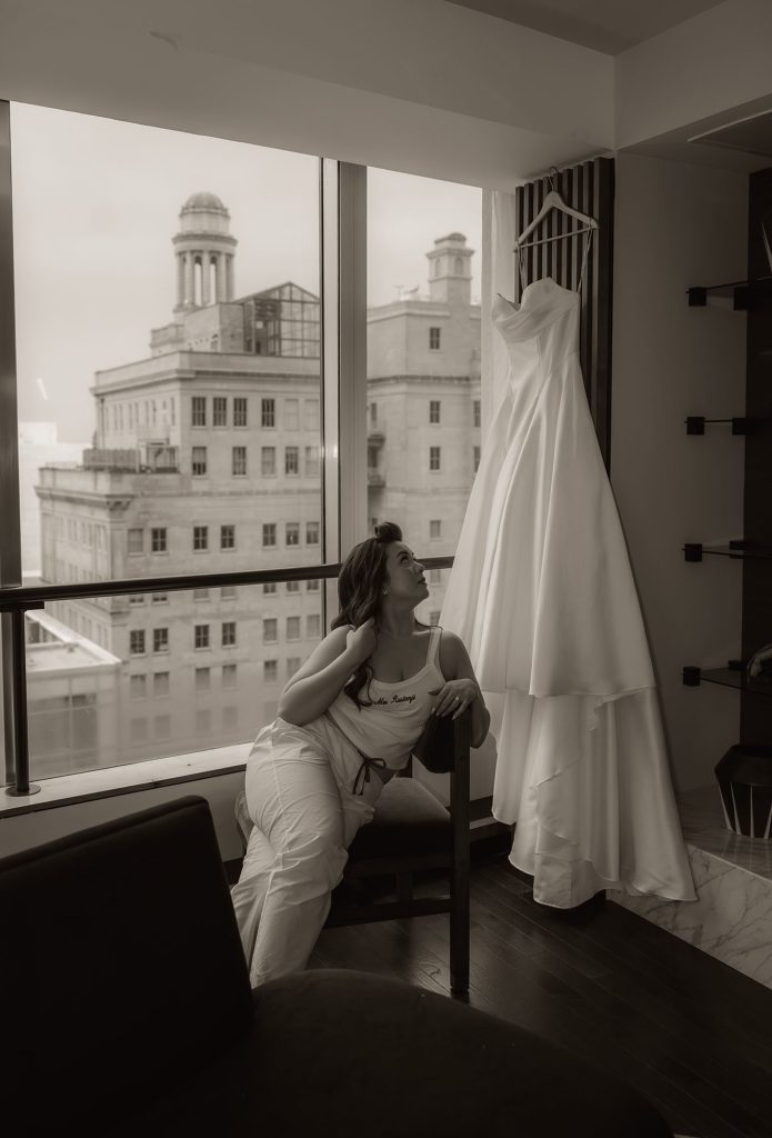Bride sitting by a large hotel window gazing at her hanging wedding gown with a classic city skyline in the background, black and white photo.