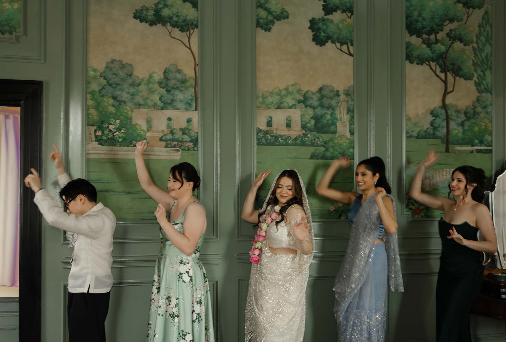 Bride in embroidered lehenga and floral garland dancing joyfully with wedding party guests in front of an elegant garden mural.